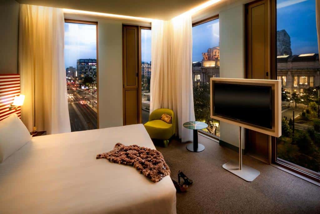 Quarto do Glam Milano com uma cama de casal, janelas panorâmicas, uma televisão e uma pequena poltrona