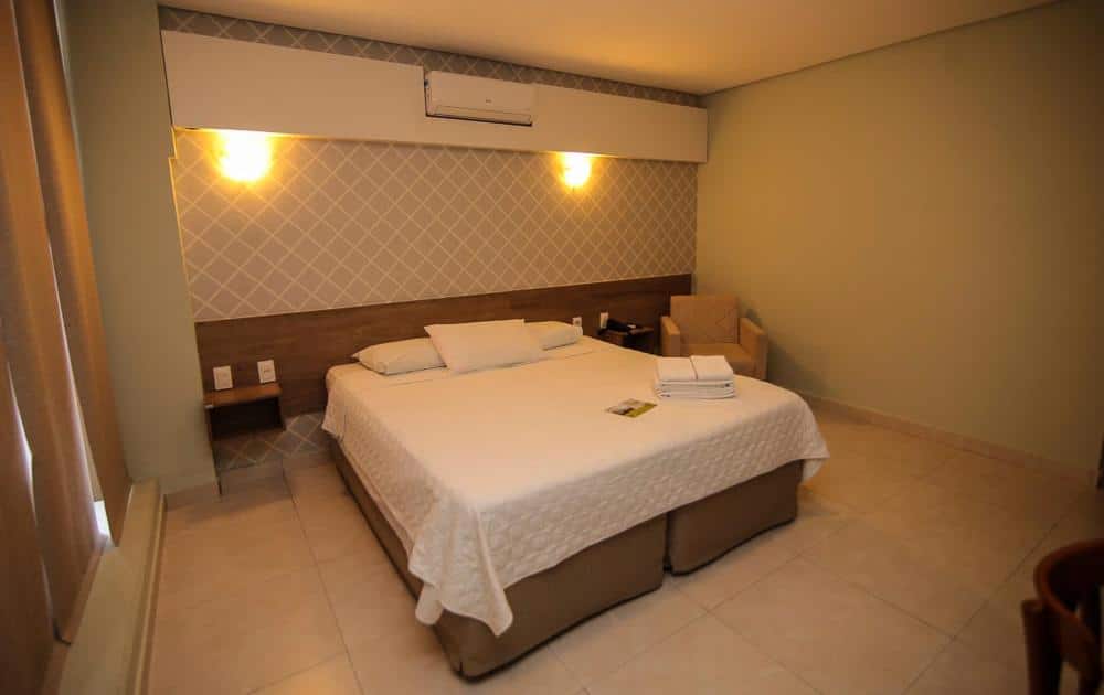 Quarto de hotel espaçoso com cama de casal, colcha branca, poltrona beje e ar-condicionado. Imagem para ilustrar o post hotéis em Teresina.