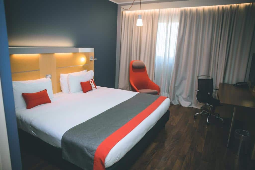 Quarto do Holiday Inn Express Milan-Malpensa Airport, an IHG Hotel com uma cama de casal, uma janela com cortinas, uma mesa de escritório com uma cadeira giratória e uma poltrona vermelha