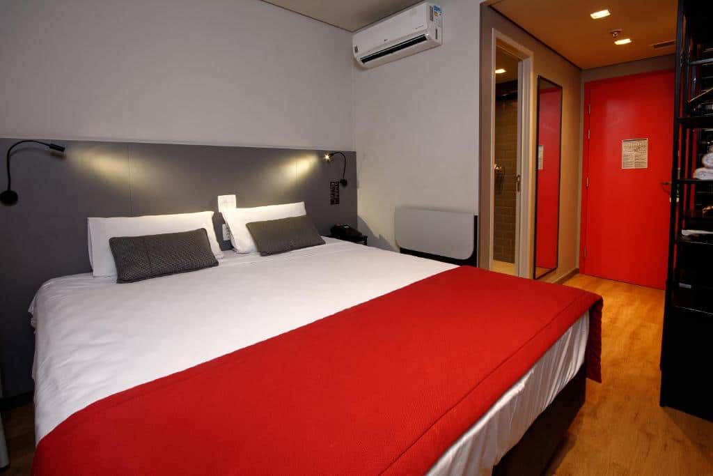 Uma cama de casal, com um ar-condicionado e um corredor na lateral. Foto para ilustrar post sobre hotéis perto do Hospital Albert Einstein.