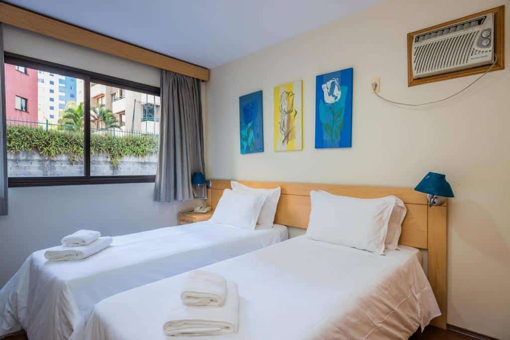 Duas camas de solteiro e um ar-condicionado em cima. Uma janela grande no quarto. Foto para ilustrar post sobre Hotéis perto do Hospital Sírio Libanês.