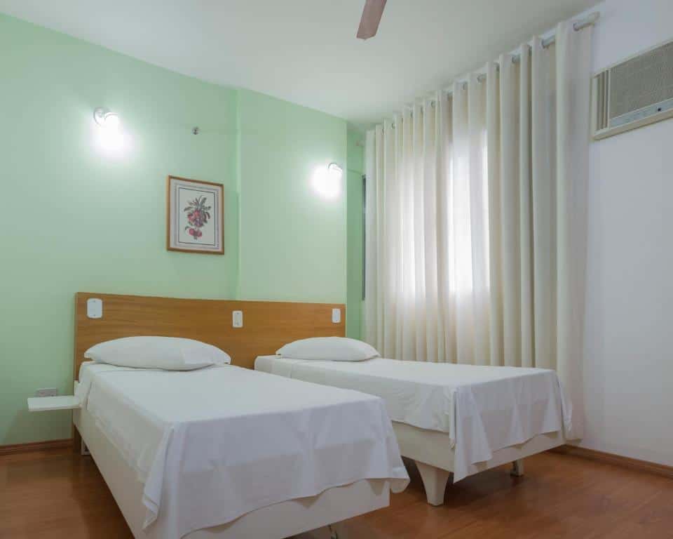 Duas camas de solteiro, atrás uma parede na cor verde claro. Uma janela com cortina. Foto para ilustrar post sobre Hotéis perto do Hospital Sírio Libanês.