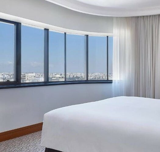 Uma televisão no painel no lado esquerdo. No lado direito uma cama de casal no Renaissance São Paulo Hotel. De frente uma grande janela com vista para a cidade. Foto para ilustrar post sobre Hotéis perto do Hospital Sírio Libanês.