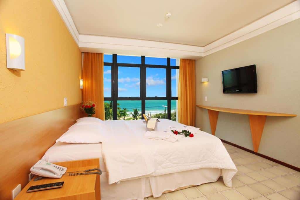 Uma cômoda com telefone, atrás uma cama e uma parede com janelas com vista para o mar. No lado direito uma televisão. Foto para ilustrar post sobre hotéis perto do Consulado Americano em Recife.