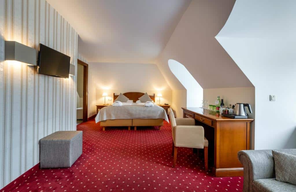 Quarto do hotel com cama de casal, carpete vermelho no chão, mesinha de madeira com poltrona marrom, tv na parede com detalhes listrados e um sofá no canto.