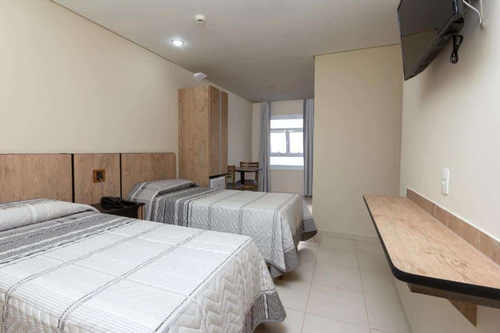 Quarto do hotel com duas camas, móveis de madeira, tv na parede, armário, uma mesa com duas cadeiras em frente a janela de vidro.
