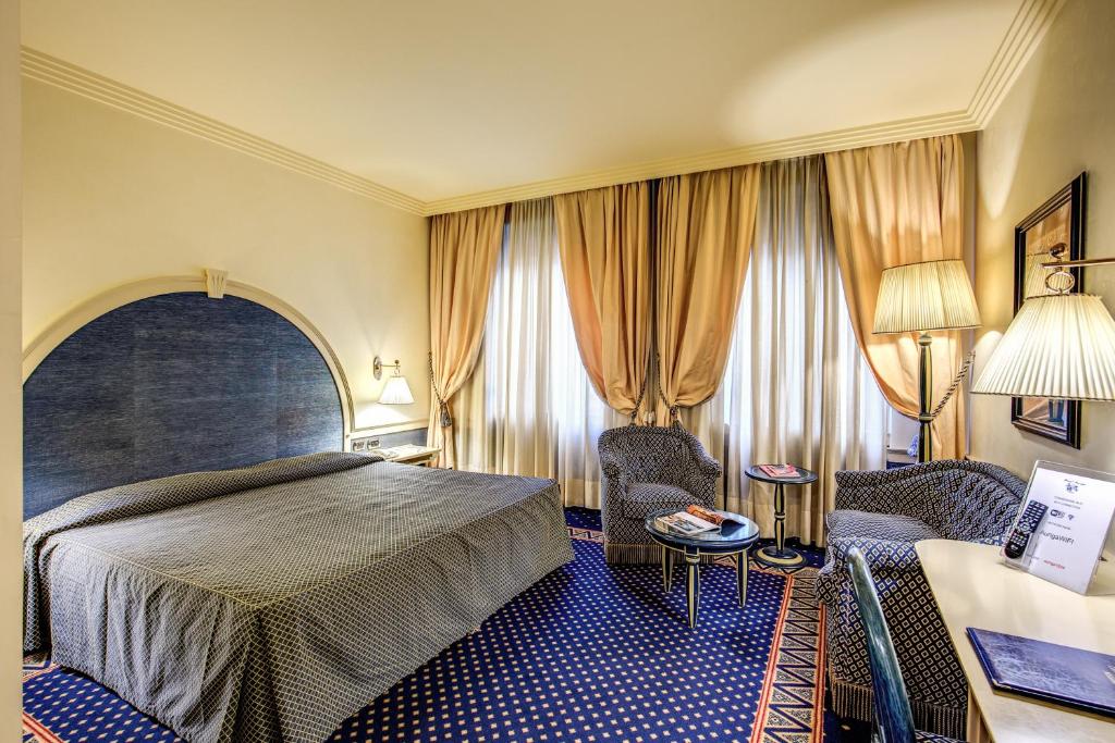 Quarto do Hotel Auriga com uma cama de casal, duas janelas amplas com cortinas, um sofá com dois lugares e um poltrona, todos os itens são estofadas em tons de branco e azul, e o restante da decoração é bege, para representar hotéis perto da Estação Central em Milão