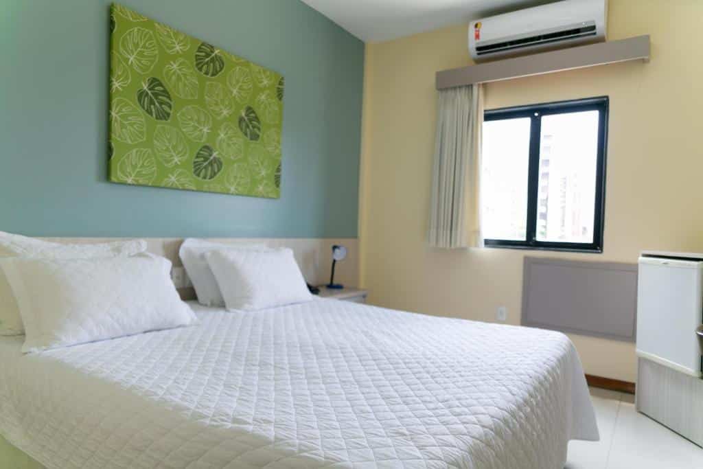Quarto de hotel com paredes azul e amarelo claro, quadro decorativo verde, cama de casal, janela e ar-condicionado. Imagem para ilustrar o post hotéis em Teresina.