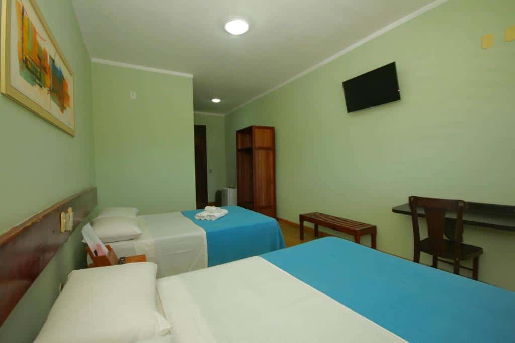Quarto de hotel com duas camas de casal, decoração simples, paredes em um tom de verde claro e móveis em madeira escura. Imagem para ilustrar o post hotéis em Parnaíba.