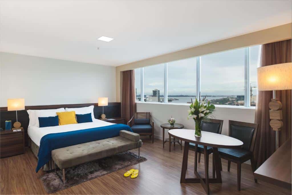 Quarto no Golden Tulip Porto Vitória com uma cama de casal, recamier, cadeiras e uma mesa com vaso de flores. Há também abajures e uma grande janela com cortinas e vista para a cidade, para representar hotéis em Vitória.
