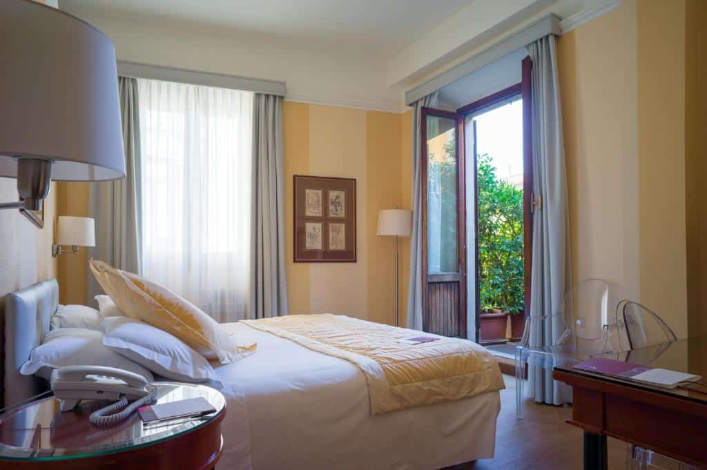 Quarto do Hotel Gran Duca Di York com uma varanda com alguns vasos de flores, há uma cama de casal, um abajur de chão, uma mesa de escritório e duas cadeiras, para representar hotéis perto do Duomo em Milão