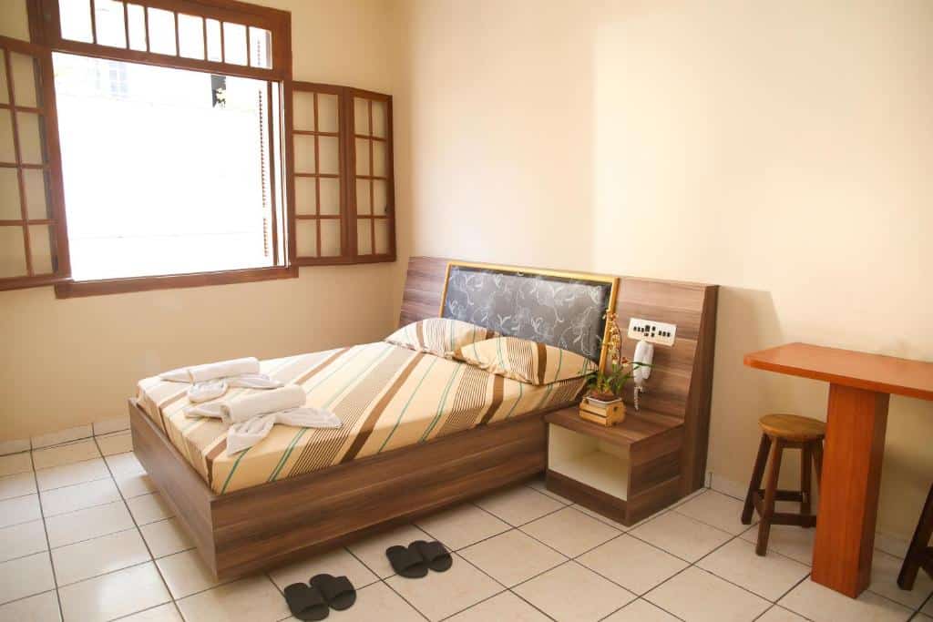 Quarto do hotel com uma cama de casal, duas toalhas em cima, dois chinelos pretos, móveis de madeira e uma janela aberta.