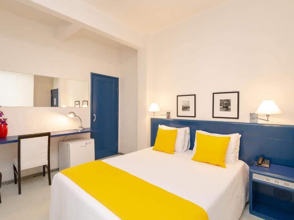 Quarto do hotel com uma cama de casal branca e amarela, móveis azuis, dois quadros na parede, espelho na parede, frigobar e uma mesa com cadeira.