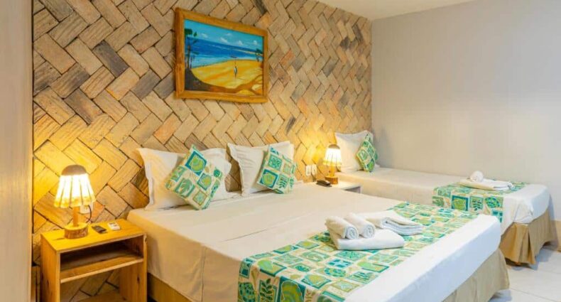 Quarto de hotel com uma cama de casal, uma cama de solteiro, decoração em tons de marrom, branco e verde, luminárias amarelas e um quadro decorativo. Imagem para ilustrar o post hotéis em Parnaíba.