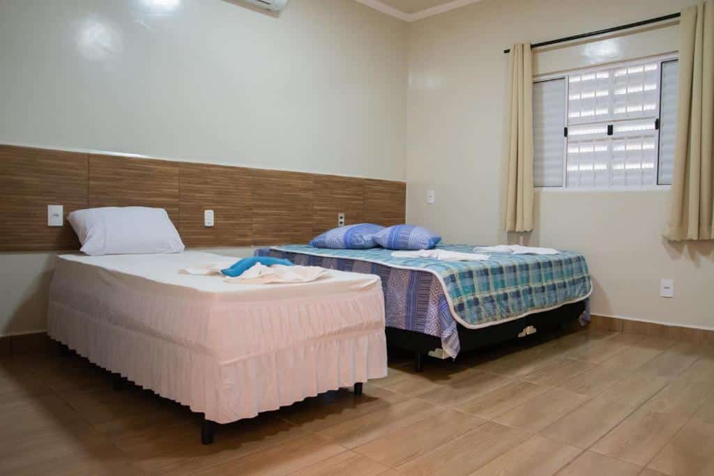 Quarto da pousada com duas camas, uma de casal e a outra de solteiro, janela branca com cortinas e detalhes de madeira na parede, ilustrando post Hotéis em Barretos.