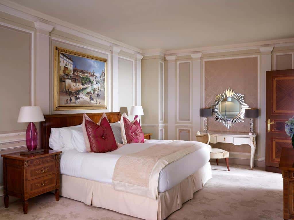 Quarto do Hotel Principe Di Savoia - Dorchester Collection em estilo renascentista, em tons de branco e rosa claro, há uma cama de casal, uma penteadeira com um espelho redondo, dois móveis de cabeceira em madeira e um quadro sob a cama
