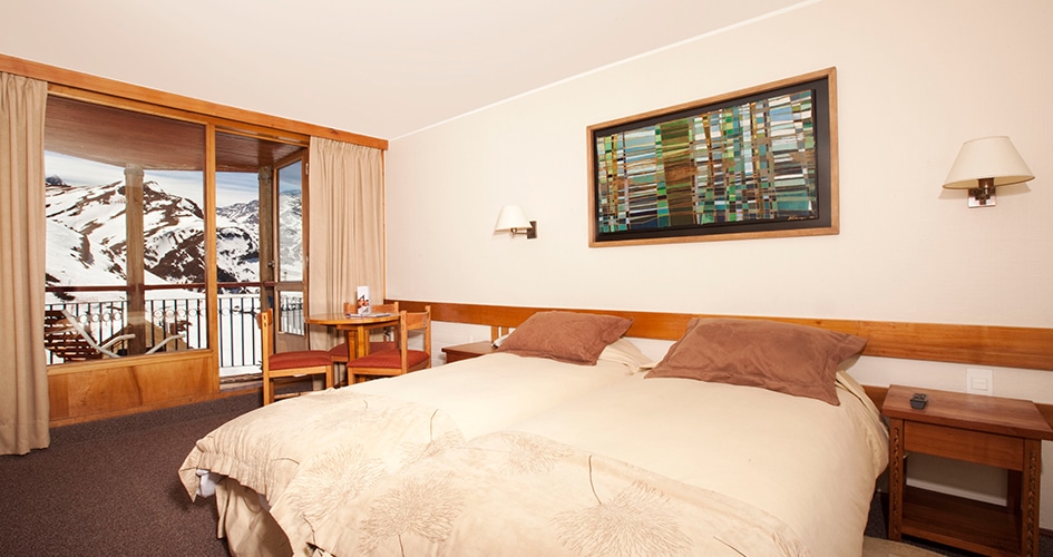 Quarto do Hotel Puerta del Sol  com cama de casal do lado direito com duas cômodas ao lado da cama. Representa hotéis no Valle Nevado.