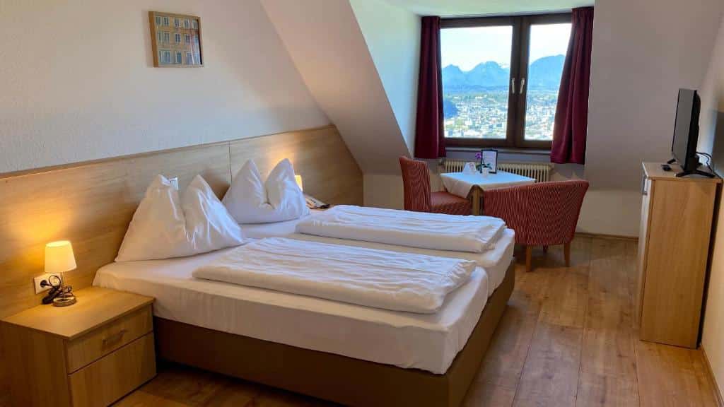 Quarto do hotel com cama de casal branca, chão e detalhes na parede de madeira, mesinha ao lado da cama, tv e duas cadeiras com uma mesa em frente a janela de vidro.
