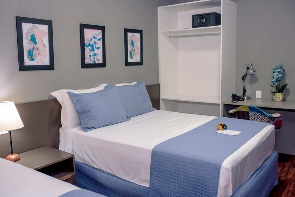 Quarto com cama de casal com colcha branca e azul, móveis brancos, parede cinza e quadros decorativos. Imagem para ilustrar o post hotéis em Teresina.