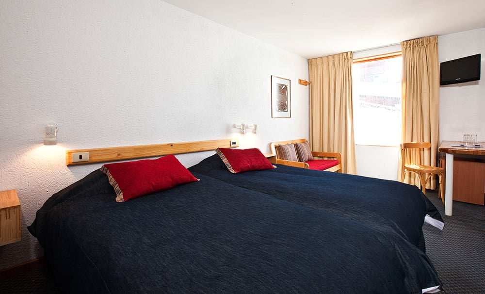 Quarto do Hotel Tres Puntas com cama de casal do lado esquerdo no centro do quarto e do lado esquerdo da cama uma poltrona com estofado vermelho. Representa onde ficar no Valle Nevado.