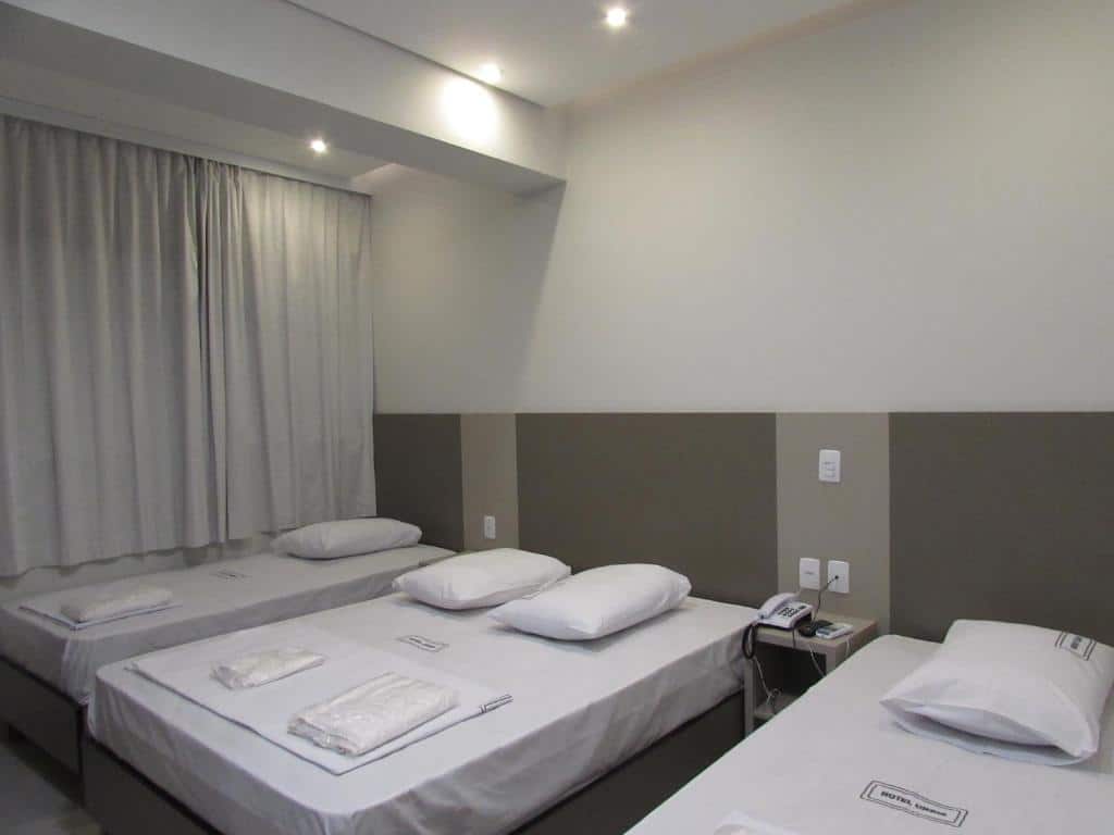 Quartos com três camas brancas, parede branca com detalhes marrom e cortina na parede.