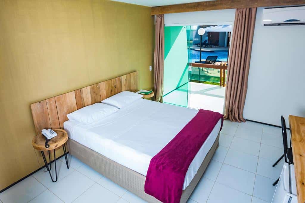Quarto de hotel com cama de casal, móveis de madeira, varanda com vista para a piscina, cortinas e ar-condicionado. Imagem para ilustrar o post hotéis em Parnaíba.