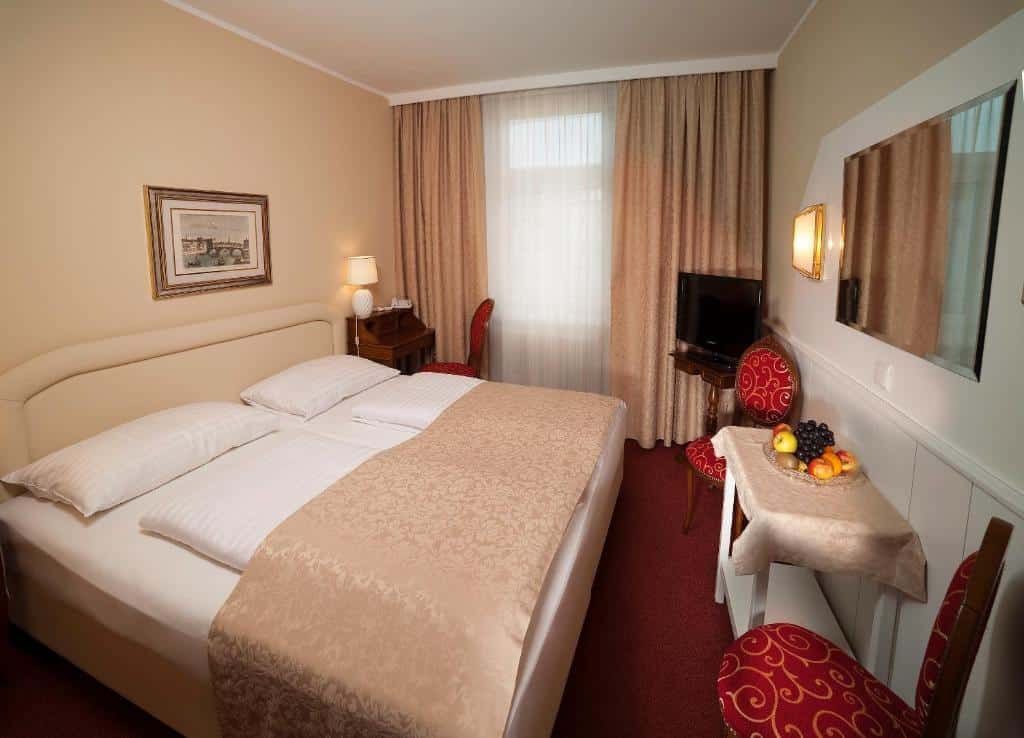 Quarto do hotel com uma cama de casal branca com detalhes em marrom, duas cadeiras vermelhas, uma mesa com frutas, quadro na parede, tv e uma janela com cortinas.