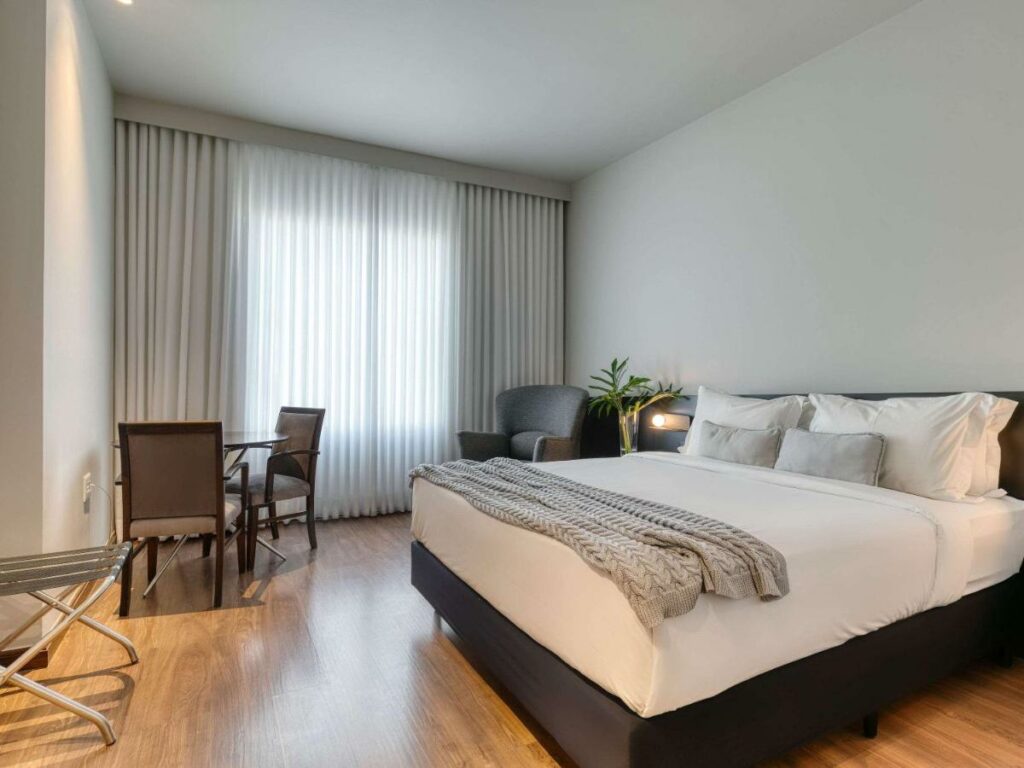 Quarto de hotel com cama de casal com lençóis brancos, janela com cortina e mesa abaixo da janela com abajour e mesa de canto.