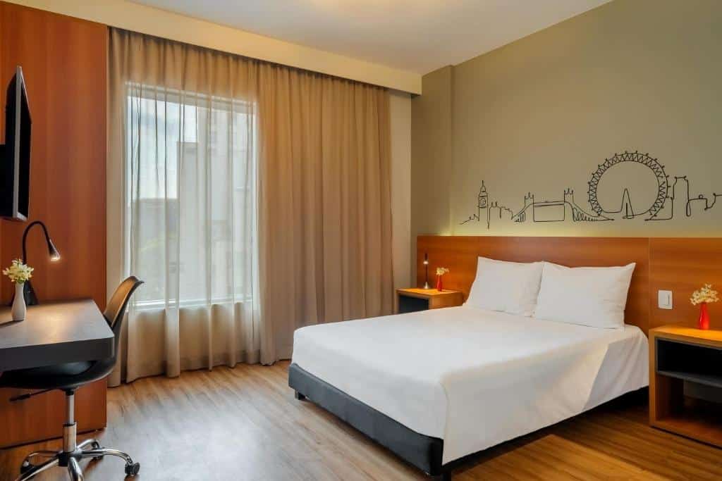 Quarto do hotel com uma cama de casal, móveis de madeira, tv na parede, desenhos na parede atrás da cama e uma janela de vidro com cortina.