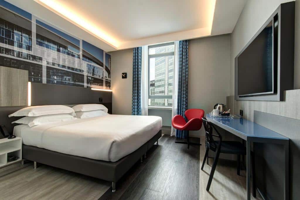Quarto no iQ Hotel Milano com uma janela com cortinas, uma cama de casal, uma mesa de escritório com uma cadeira, uma televisão e uma poltrona vermelha, para representar os melhores hotéis em Milão