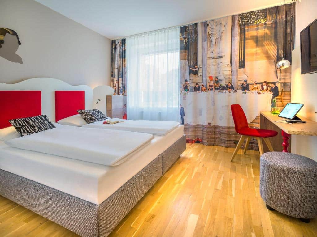 Quarto do hotel com uma cama de casal branca com detalhes em vermelho, tv na parede, mesa de madeira com uma cadeira vermelha e cortina colorida.