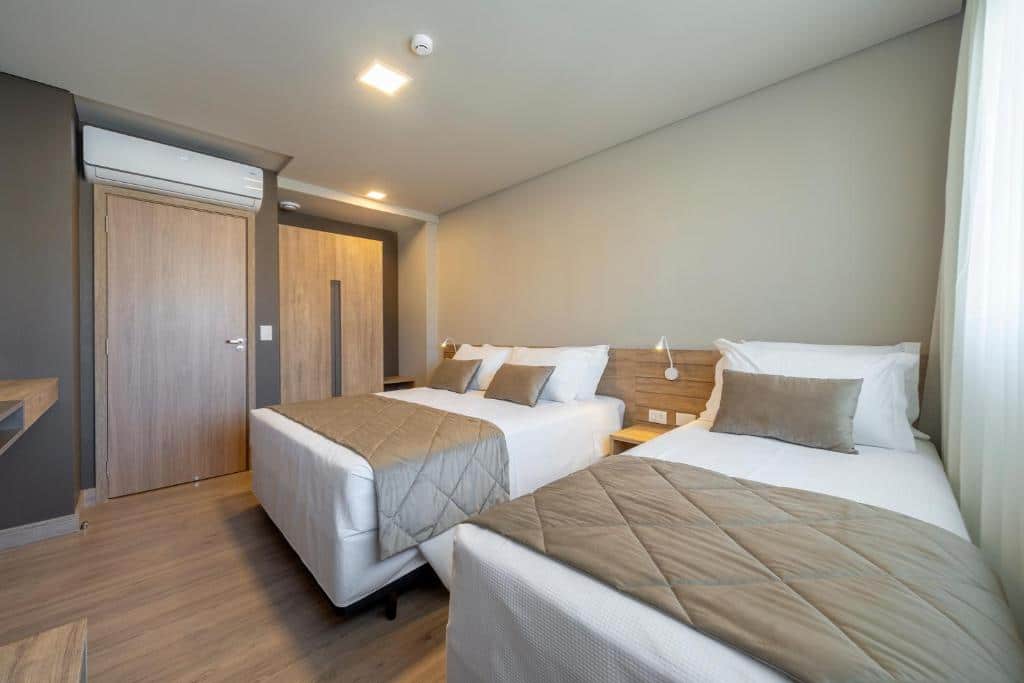 Suíte do Laghetto Resort Golden Gramado com duas camas, uma de casal e outra de solteiro, ambas situadas à direita do quarto. Ao fundo, no canto esquerdo, há uma porta larga de madeira com um ar-condicionado branco em cima da passagem.