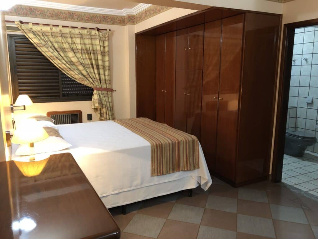 quarto do Mabruk Barretos Apart Hotel mostrando uma cama de casal à esquerda da imagem, com um grande armário de madeira em frente aos pés da cama e uma porta mostrando o banheiro à direita