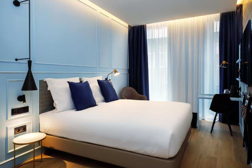 Quarto do Mercure Porto Centro Aliados com cama de casal do lado esquerdo e do lado esquerdo da cama uma pequena mesa de trabalho com cadeira. Representa hotéis Mercure no Porto.