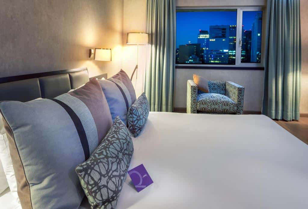 Quarto do Mercure Santiago Centro com cama de casal do lado esquerdo e do lado direito uma poltrona com janela ao fundo com cortinas. Representa hotéis Mercure em Santiago.