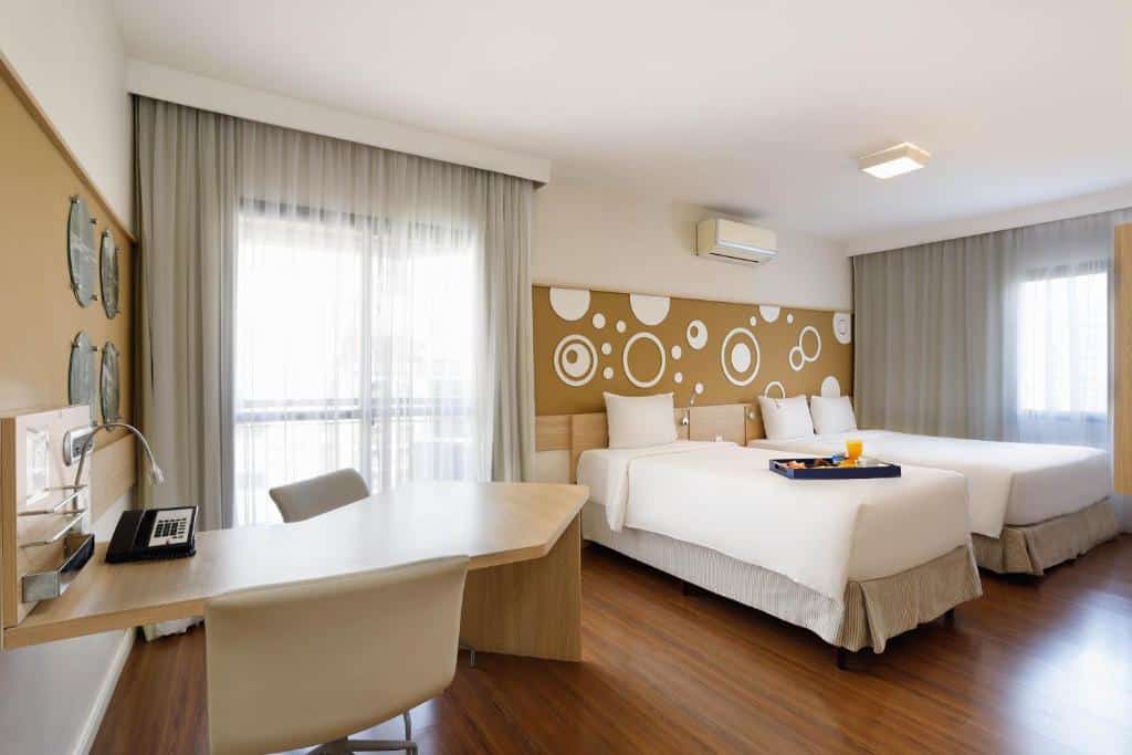 Quarto do hotel com duas camas de solteiro, desenhos de círculos atrás, ar condicionado, escrivaninha de madeira com uma cadeira e sacada de vidro com cortina.