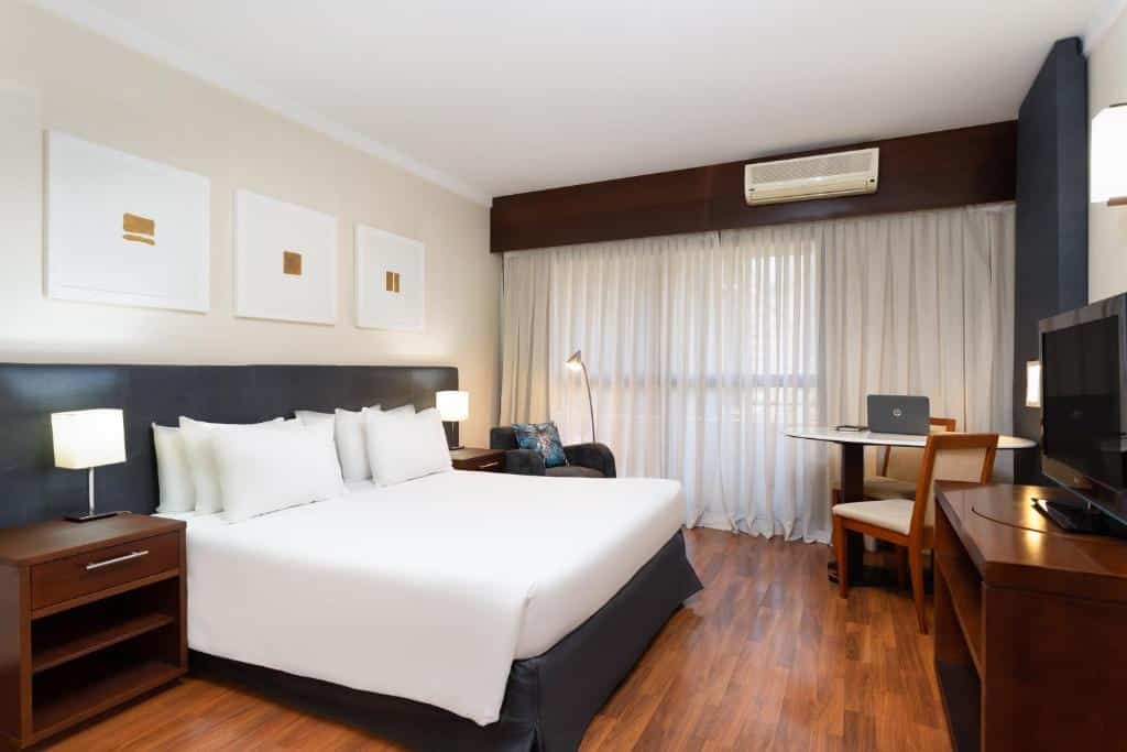 Quarto do hotel com uma cama de casal, piso e móveis de madeira, quadros na parede, mesinha ao lado da cama, tv, ar condicionado, janela com cortina e uma mesa.
