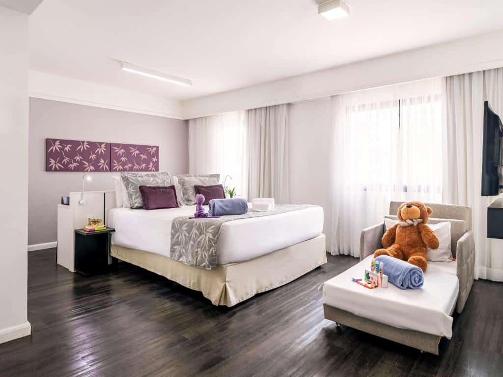 Quarto do hotel com três camas, uma de casal e uma para criança com um urso de pelúcia em cima, quadros na parede, duas janelas com cortinas e uma tv.