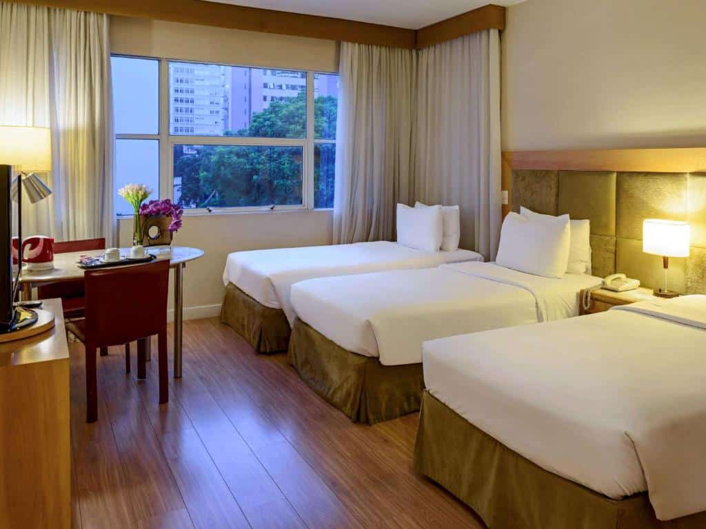 Quarto do hotel com três camas de solteiro brancas, uma tv, uma mesinha com duas cadeiras vermelhas e uma janela de vidro.