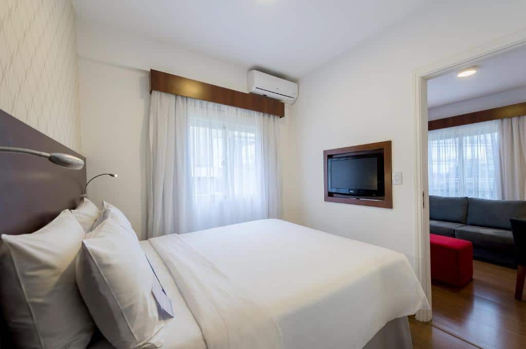 Quarto do hotel com paredes brancas, uma cama de casal branca, janela com cortina, tv na parede e porta que da acesso a sala com sofá.
