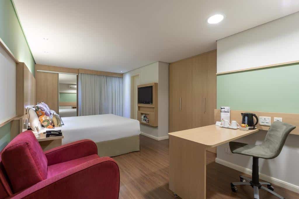 Quarto do hotel com uma cama de casal, móveis em madeira, escrivaninha com cadeira, poltrona vermelha e tv na parede.