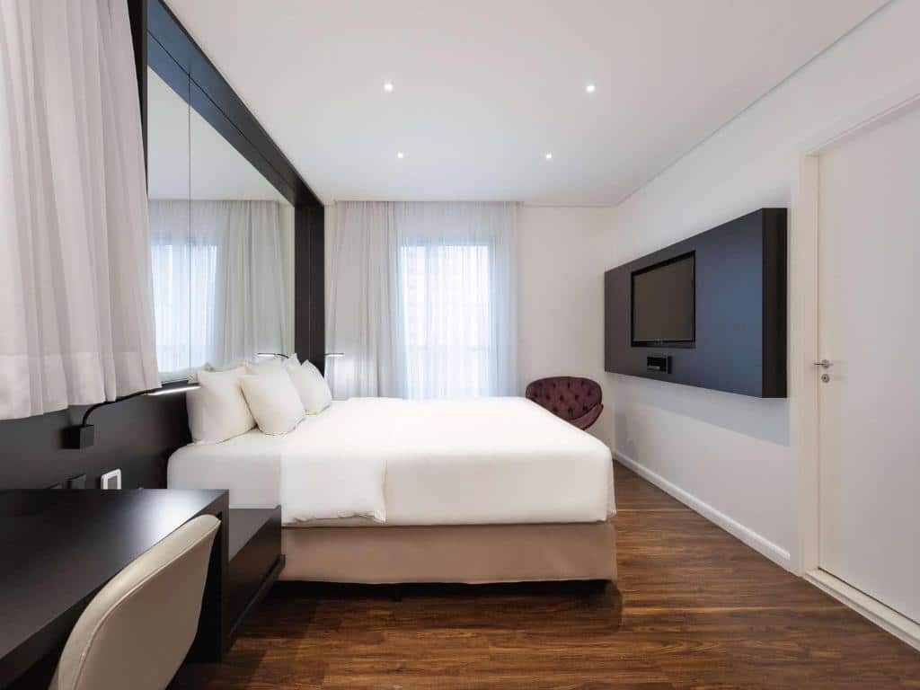 Quarto do hotel com paredes brancas, uma cama de casal, uma escrivaninha preta com cadeira, uma poltrona, tv na parede e janela com cortina branca.