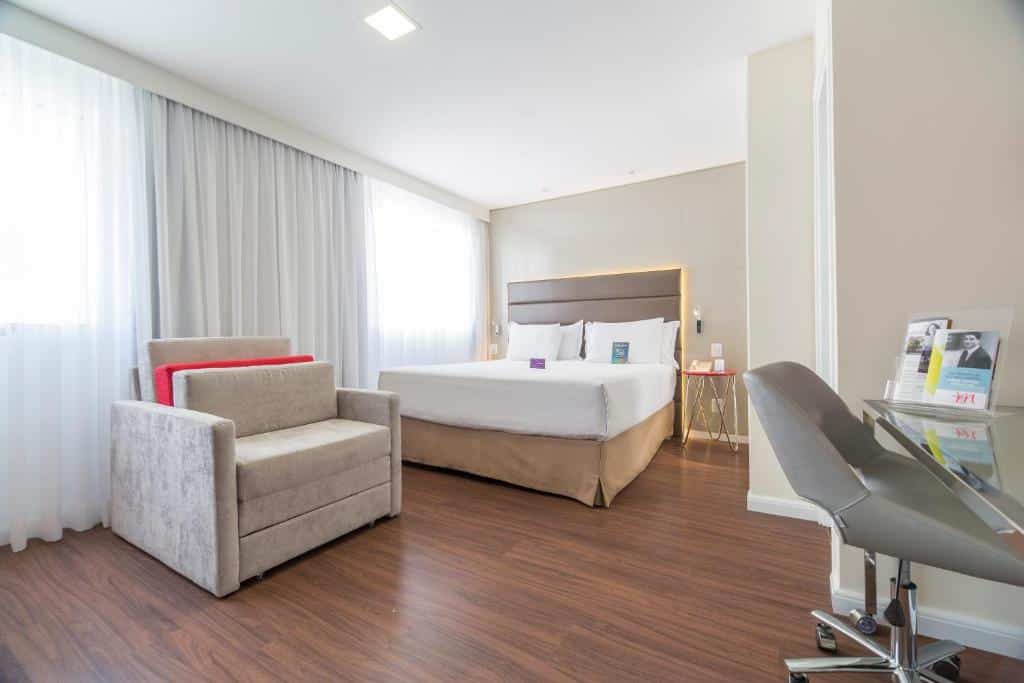 Quarto do hotel com uma cama de casal, uma cadeira marrom na escrivaninha, uma poltrona e duas janelas com cortinas brancas.