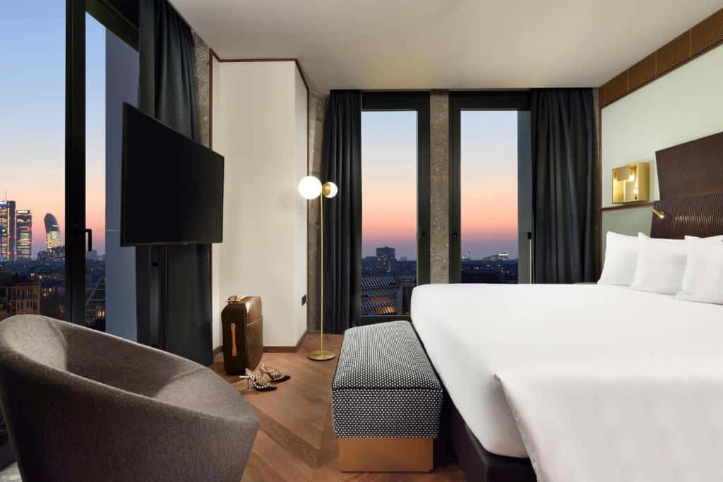 Quarto do Milano Verticale | UNA Esperienze com janelas panorâmicas, uma cama de casal, um abajur de chão, uma poltrona e uma televisão de frente para a cama, para representar hotéis de luxo em Milão