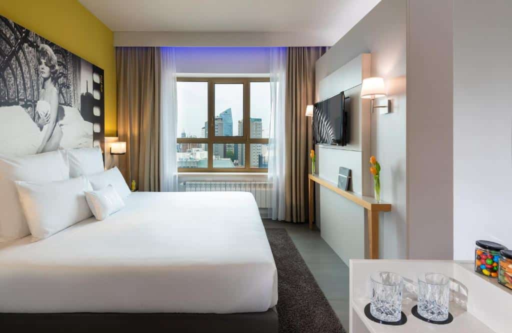 Quarto do NYX Hotel Milan by Leonardo Hotels com uma janela com cortinas, cama de casal, uma televisão e uma pequena mesa com copos e alguns petiscos doces