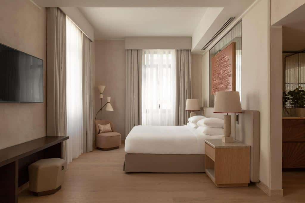 Quarto do Park Hyatt Milano com uma cama de casal, com duas janelas com cortinas, uma poltroninha com um abajur de chão, há duas mesinhas de cabeceira e uma televisão na parede de frente para a cama, para representar hotéis de luxo em Milão