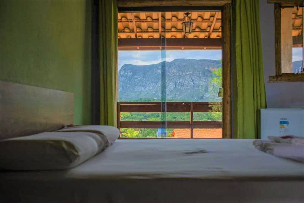 quarto da Pousada Bela Vista do Ismail em Lapinha da Serra mostrando uma cama de casal com lençóis branco em frente a uma porta de vidro que dá acesso a varanda coberta com vista para as montanhas