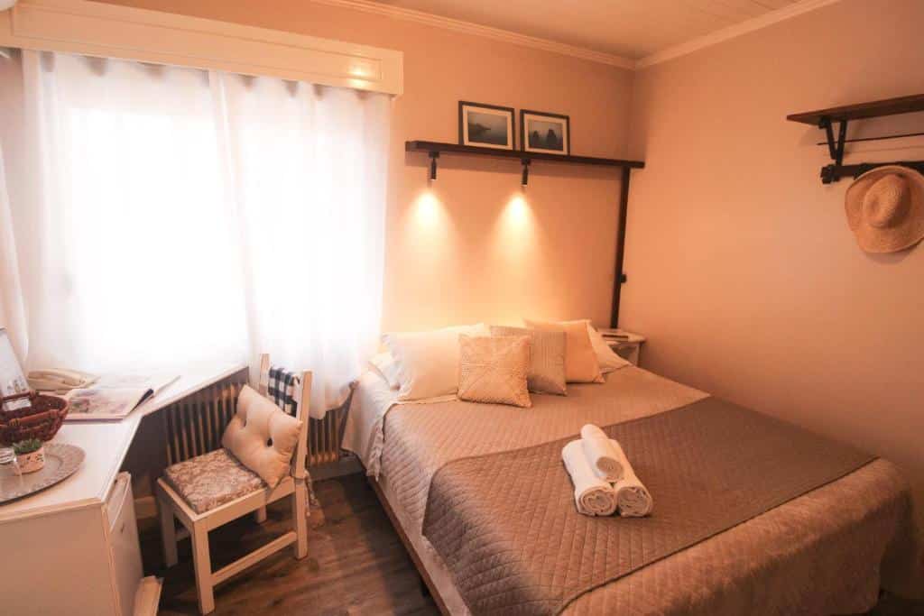 quarto da Pousada Betânia em Gramado com uma cama de casal à direita e uma mesa de canto branco com uma cadeira estofada à esquerda.