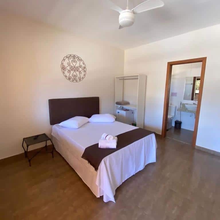 quarto da Pousada Cipó Cana na Lapinha da Serra com uma cama de casal ao centro, um pequeno armário branco ao lado e uma porta com acesso ao banheiro.
