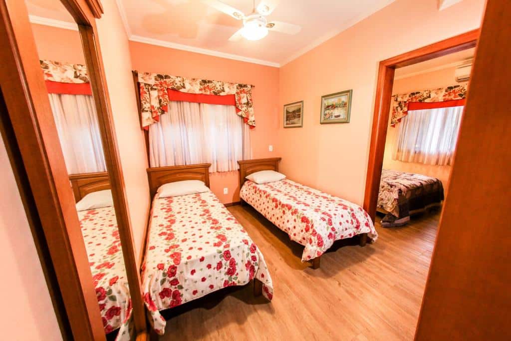 quarto da Pousada das Papoulas em Gramado com duas camas de solteiro decoradas com lençóis floridos em tons vermelhos, e uma porta à direita com acesso ao quarto ao lado mostrando uma parte de uma cama de casal
